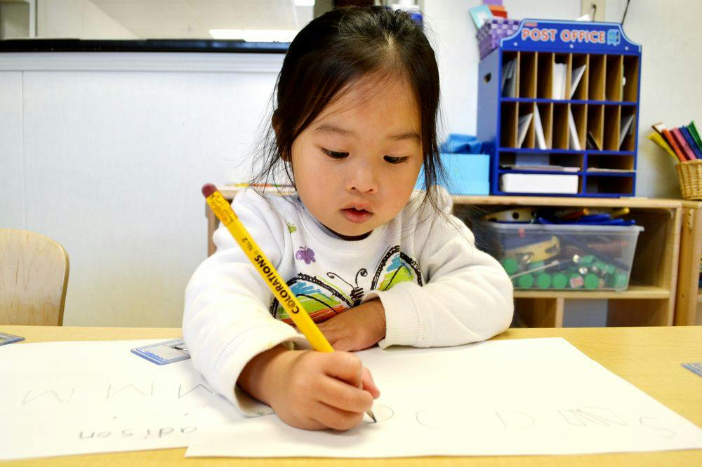 Child writing on paper with a yellow pencil in a classroom