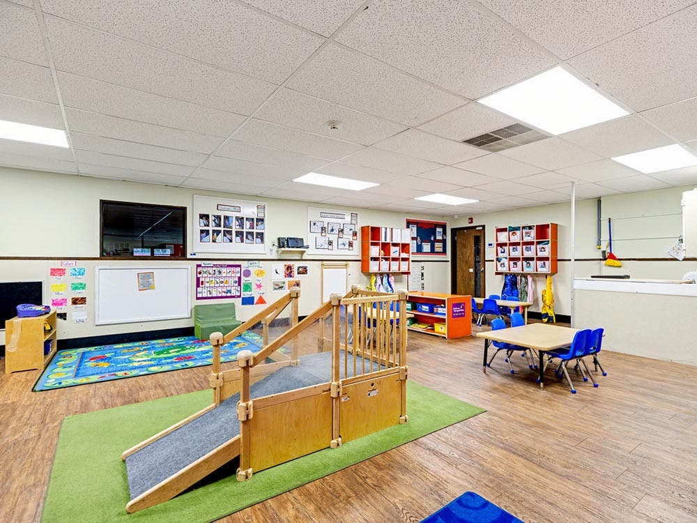 Empty classroom with small tables and chairs, play ramp, and colorful rug