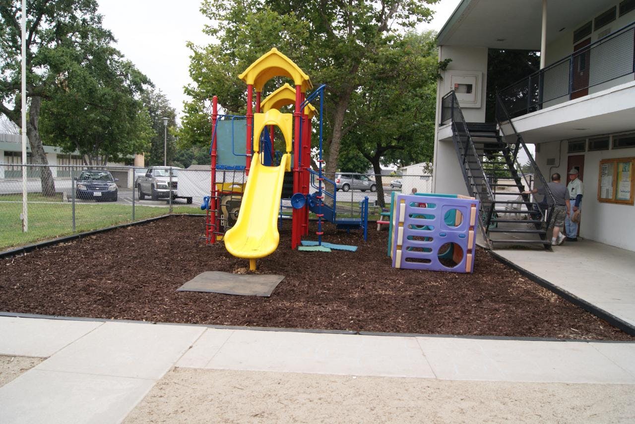 Playground with a yellow slide and climbing structure on wood chips next to a building