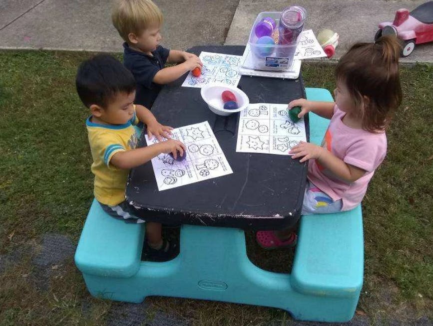 Three children coloring at a small picnic table outdoors
