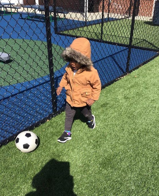 Child in a hooded jacket walking on artificial turf next to a soccer ball