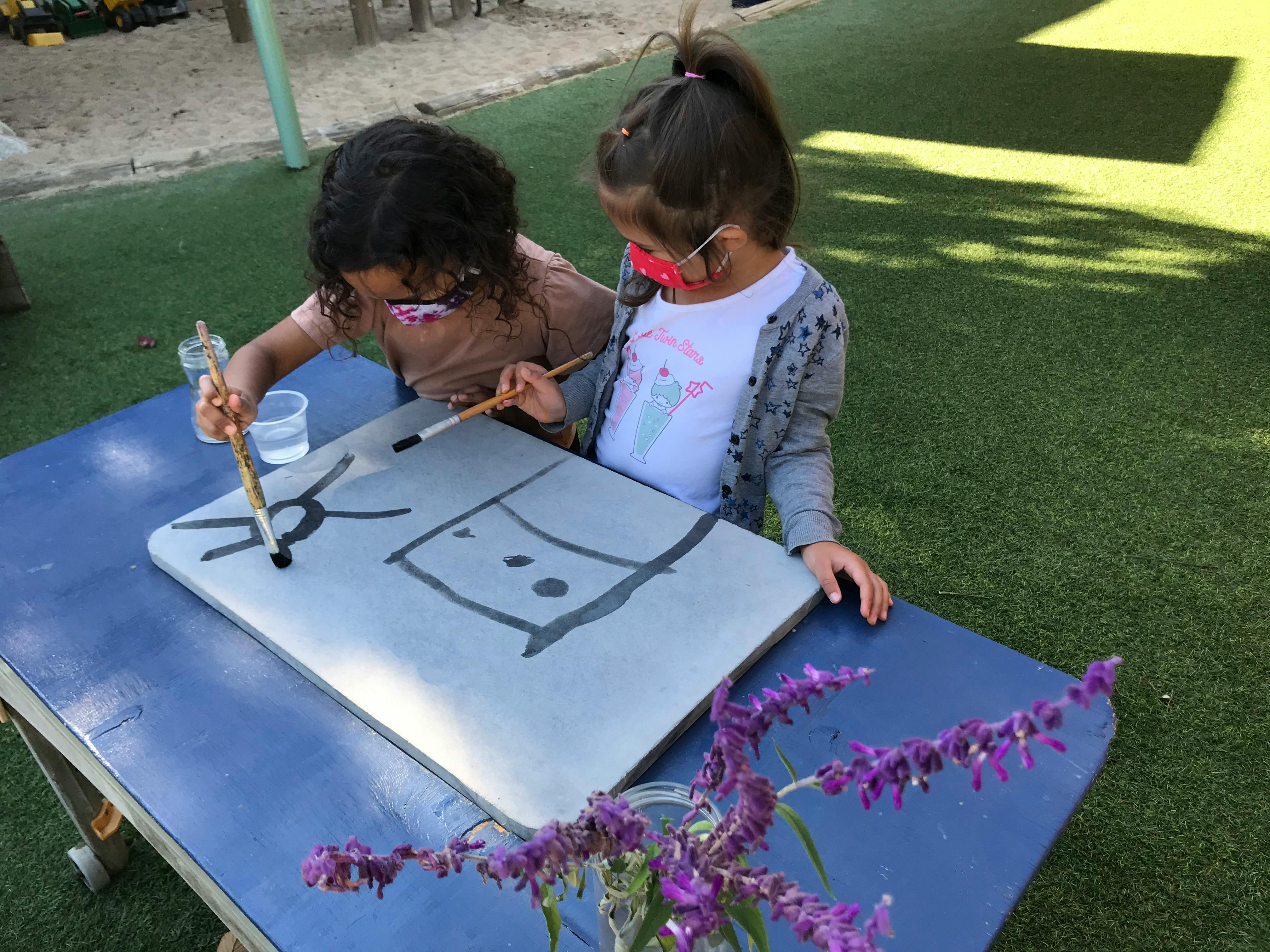Two children painting on a large white board outdoors