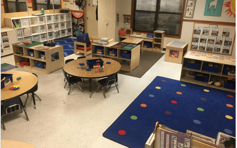 Classroom with round tables and chairs, shelves with bins, and a blue rug with colored dots