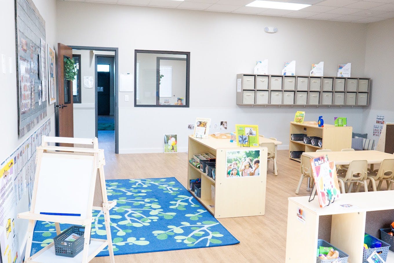 Classroom interior with small tables, chairs, shelving, and a blue area rug