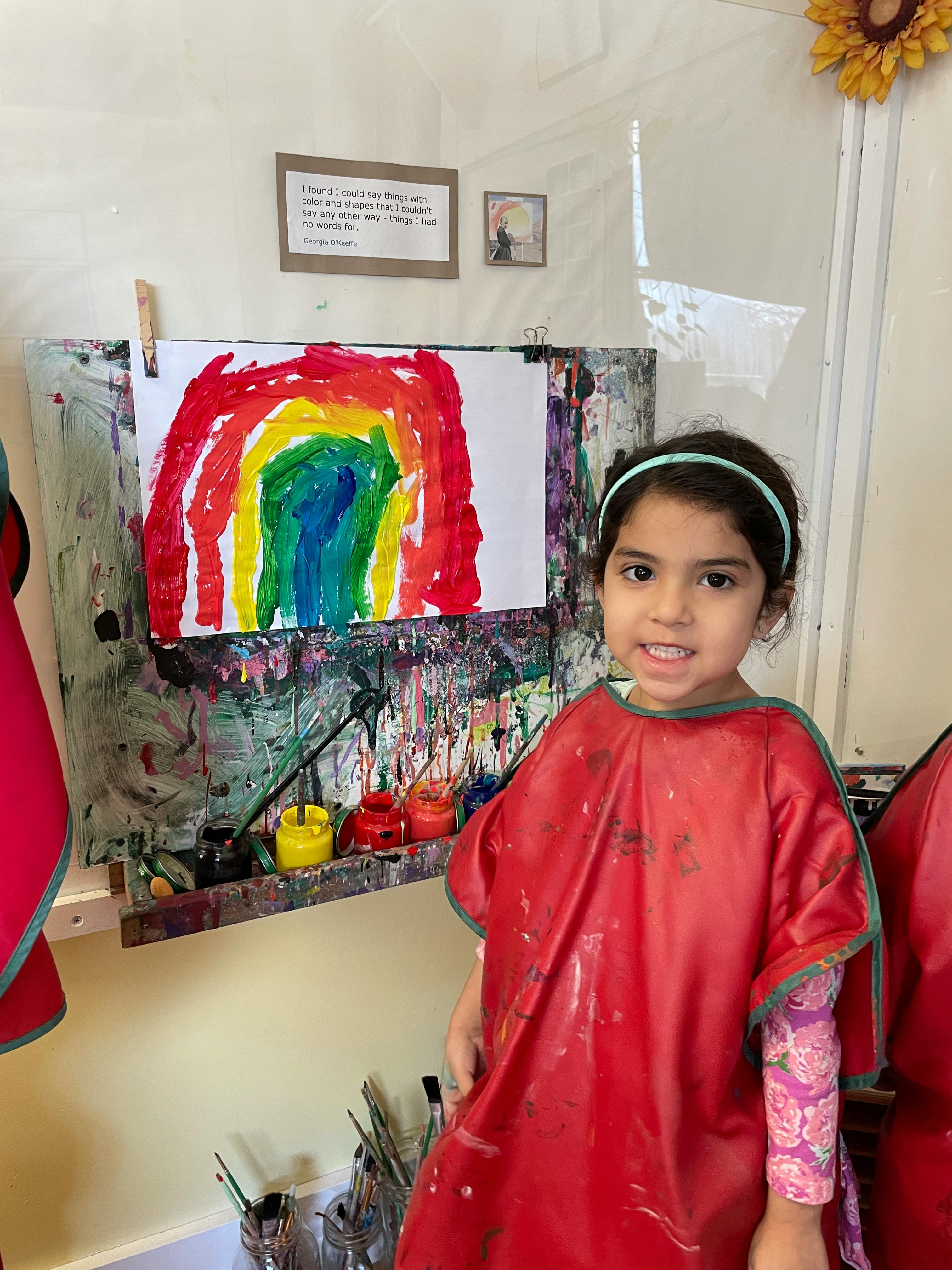 Child in a red smock standing next to a rainbow painting