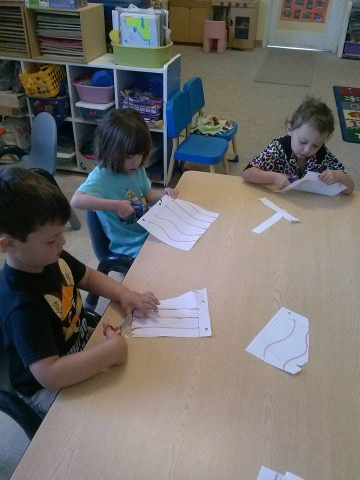 Three children cutting paper at a table in a classroom