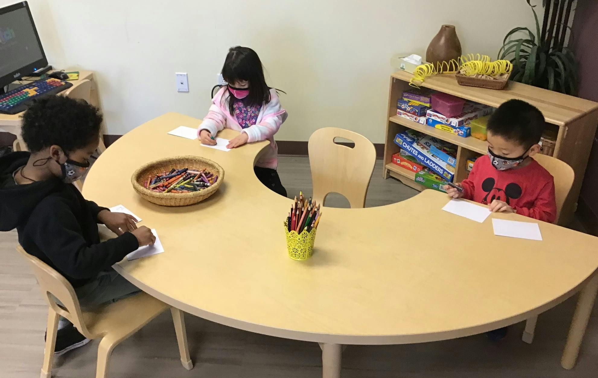 Three children drawing at a crescent-shaped table in a classroom