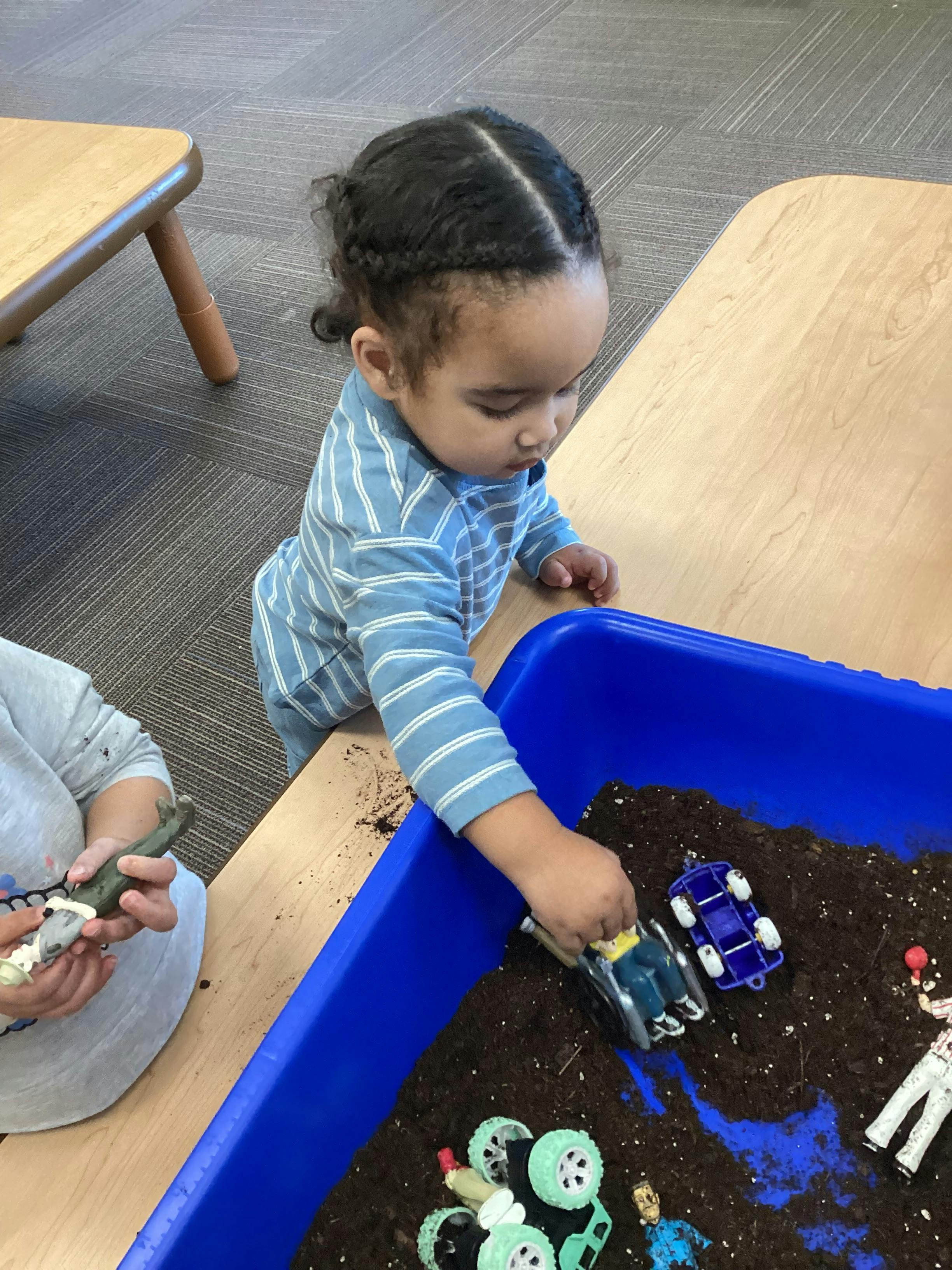 Child playing with toy cars in a blue sensory bin filled with dirt