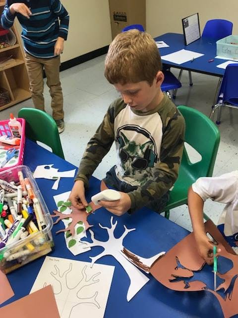 Child gluing paper leaves onto a paper tree cutout at a table