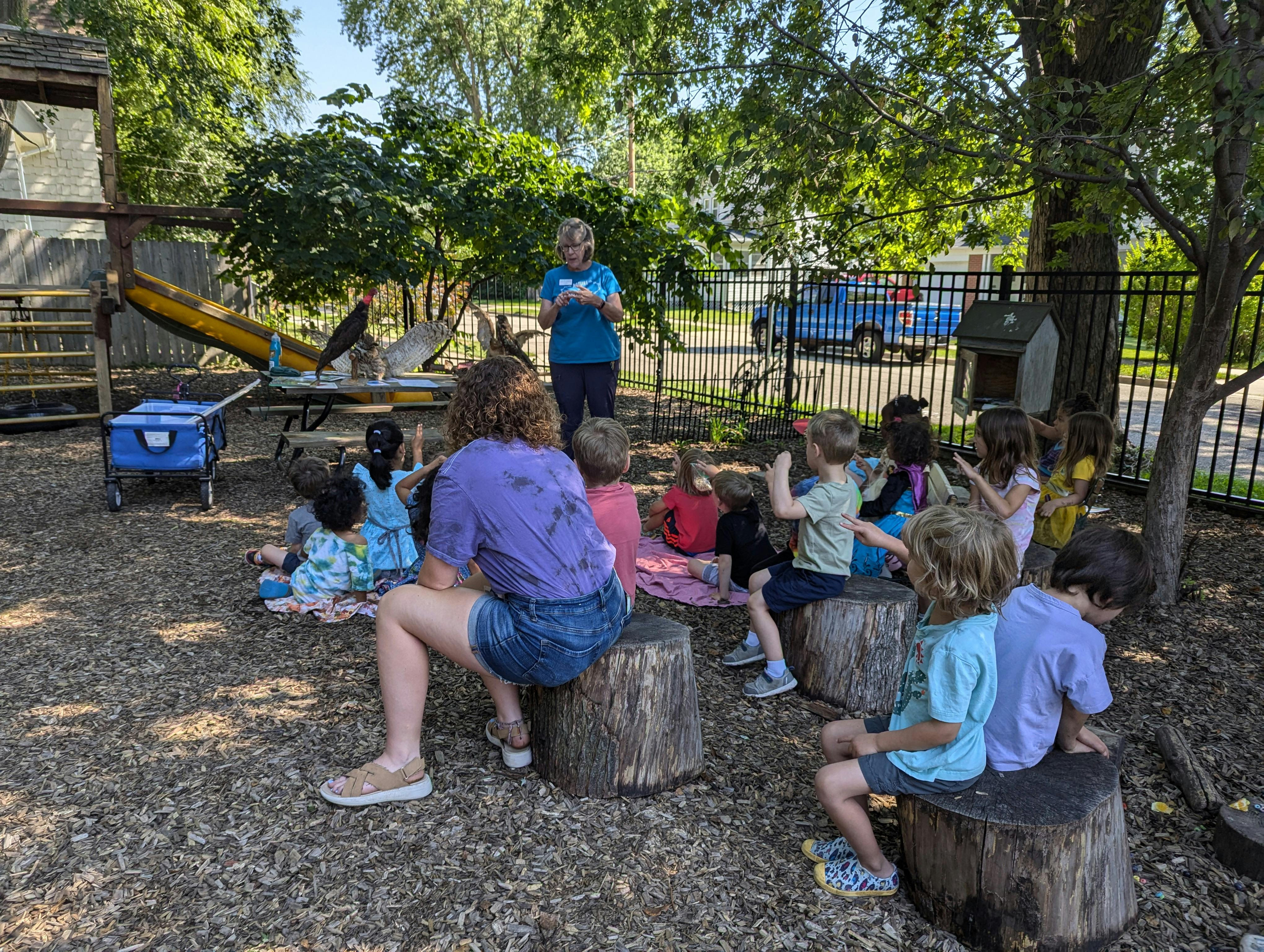 Adult speaking to a group of children sitting on tree stumps outdoors