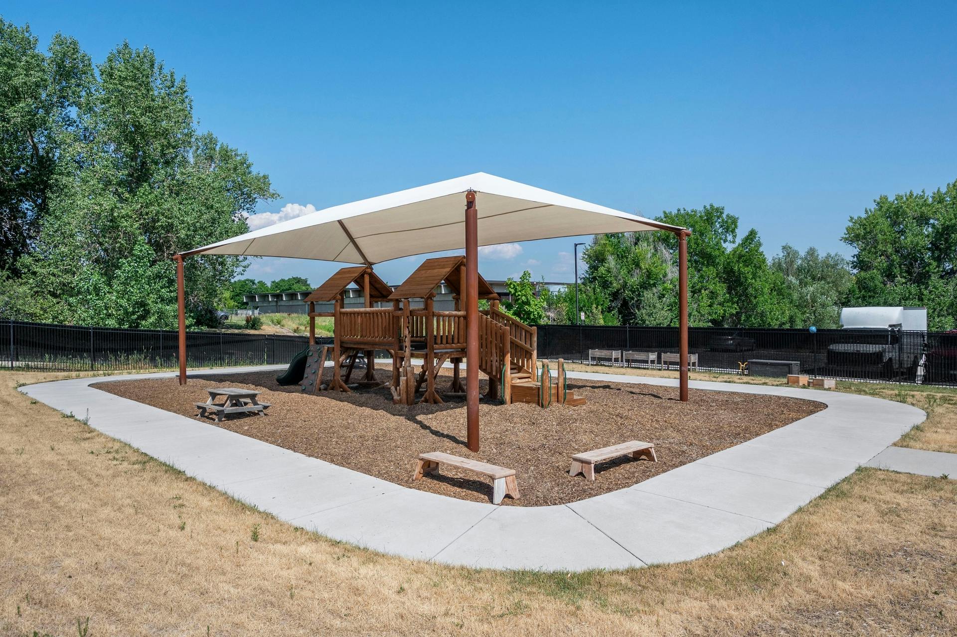 A playground with a wooden play structure, picnic table, and benches under a shade