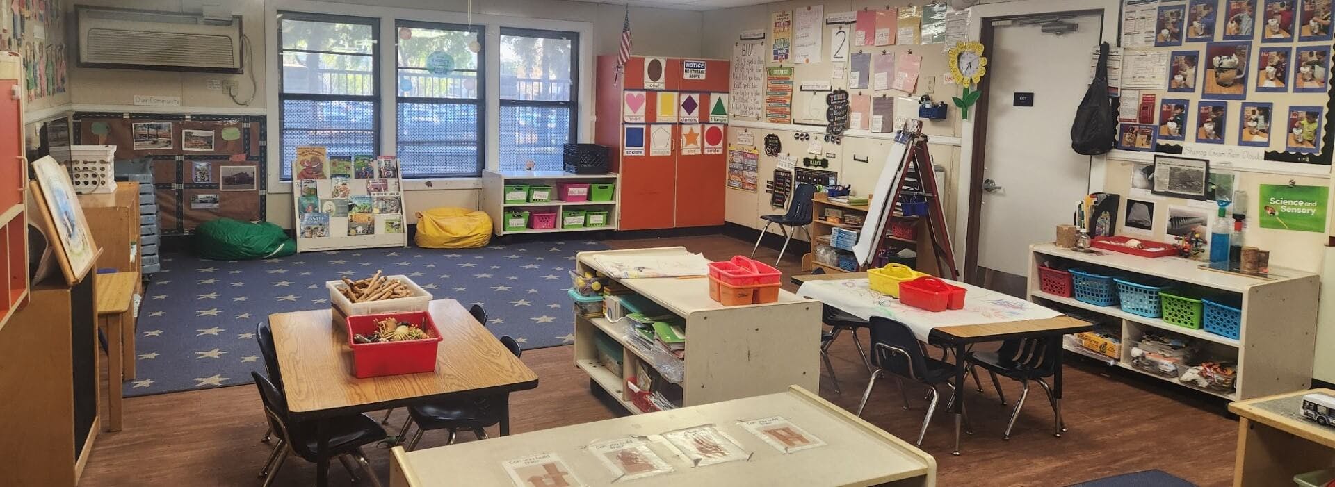 An empty classroom with small tables and chairs, shelves, and a blue rug with stars
