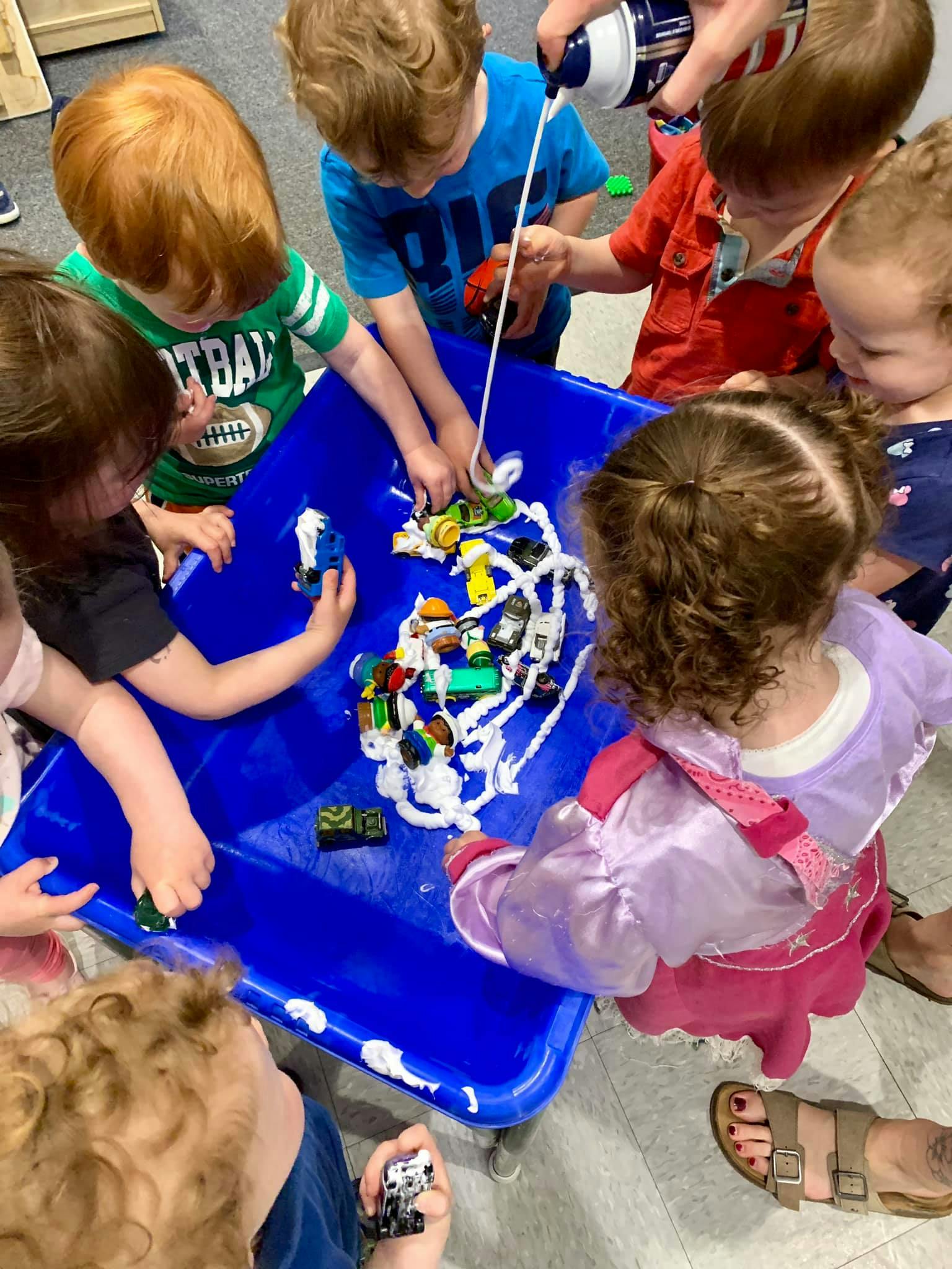 Children playing with toy cars and shaving cream in a blue bin