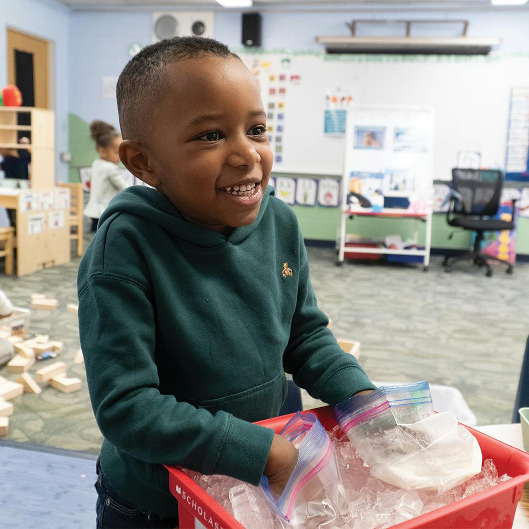 Child in a classroom holding a red bin with bags of ice