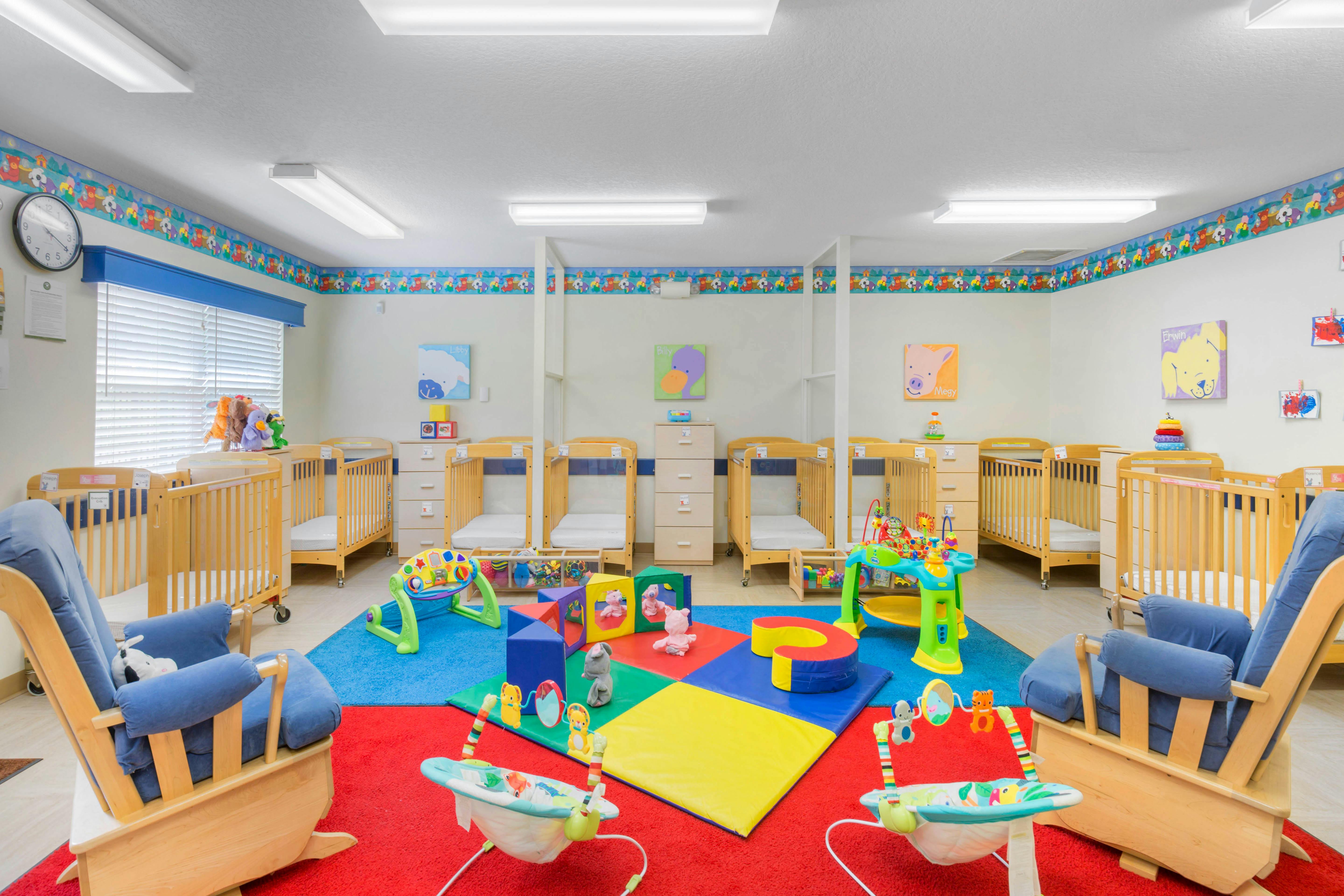 A colorful childcare room with multiple cribs, chairs, and play mats