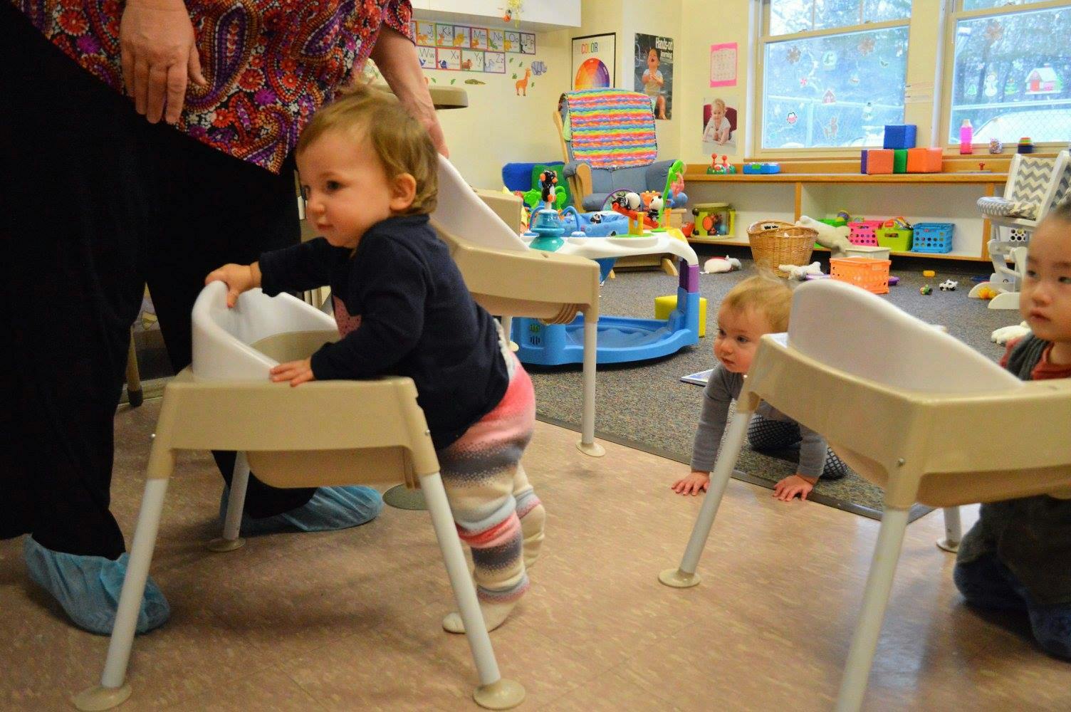 Child standing by a high chair in a classroom with other children
