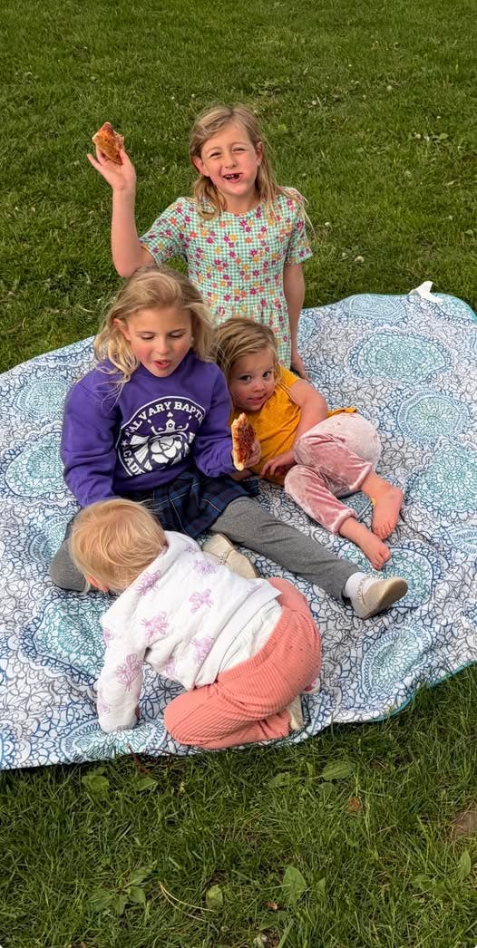 Four children sitting on a blanket on the grass, two holding pizza slices