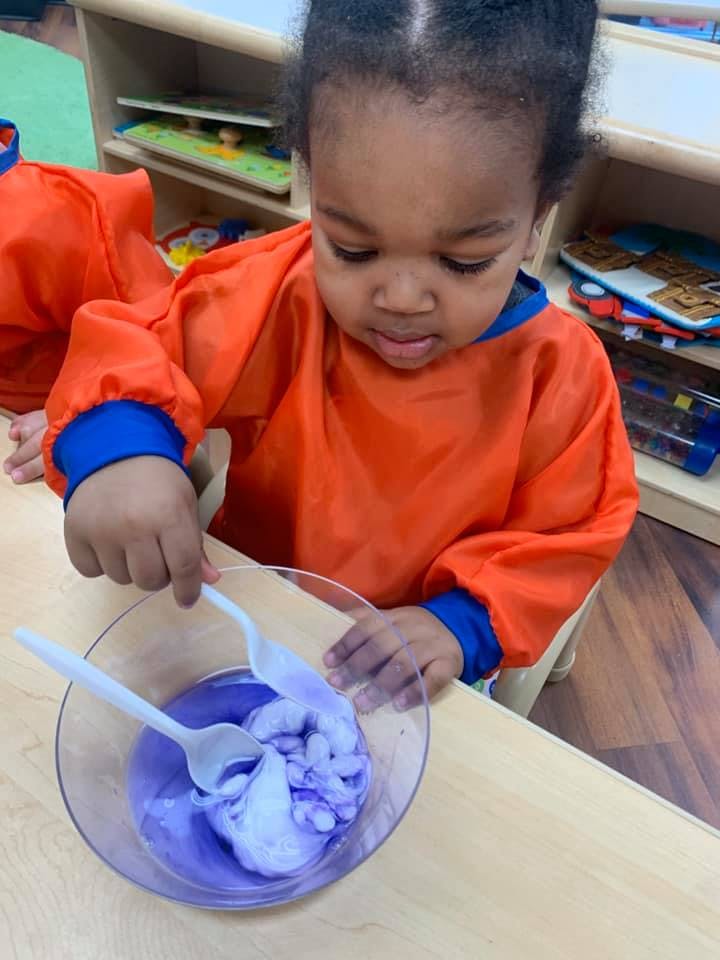 Child in orange smock mixing purple liquid in a bowl with two spoons