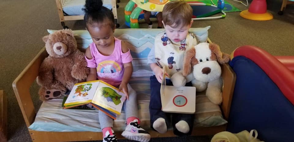 Two children sitting on a bench with stuffed animals, reading books