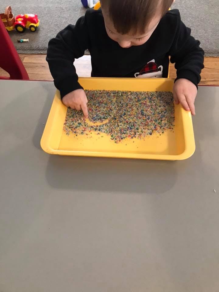 Child drawing in a tray of colorful sprinkles