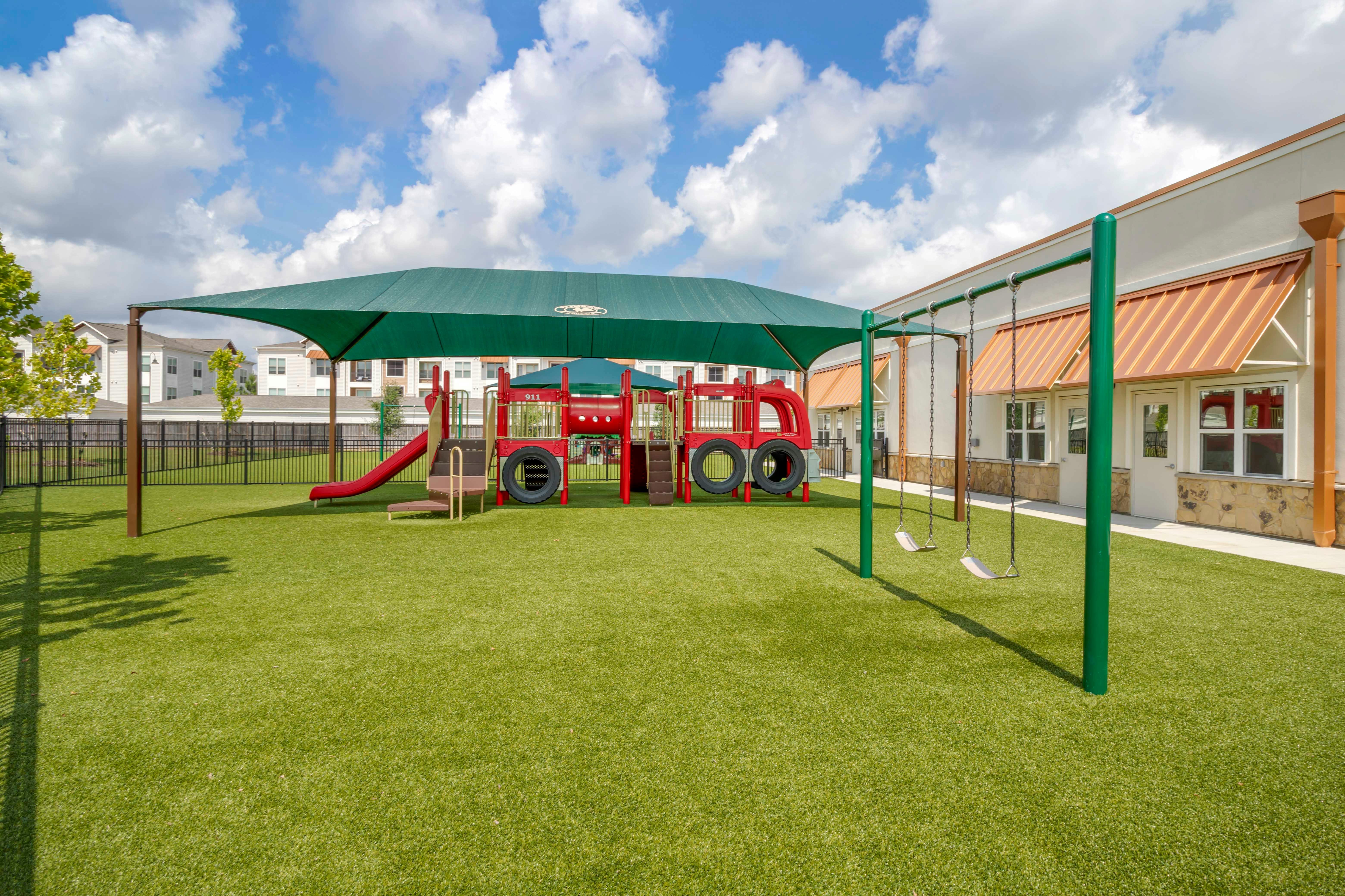 An outdoor playground with swings, a red play structure, and green artificial turf