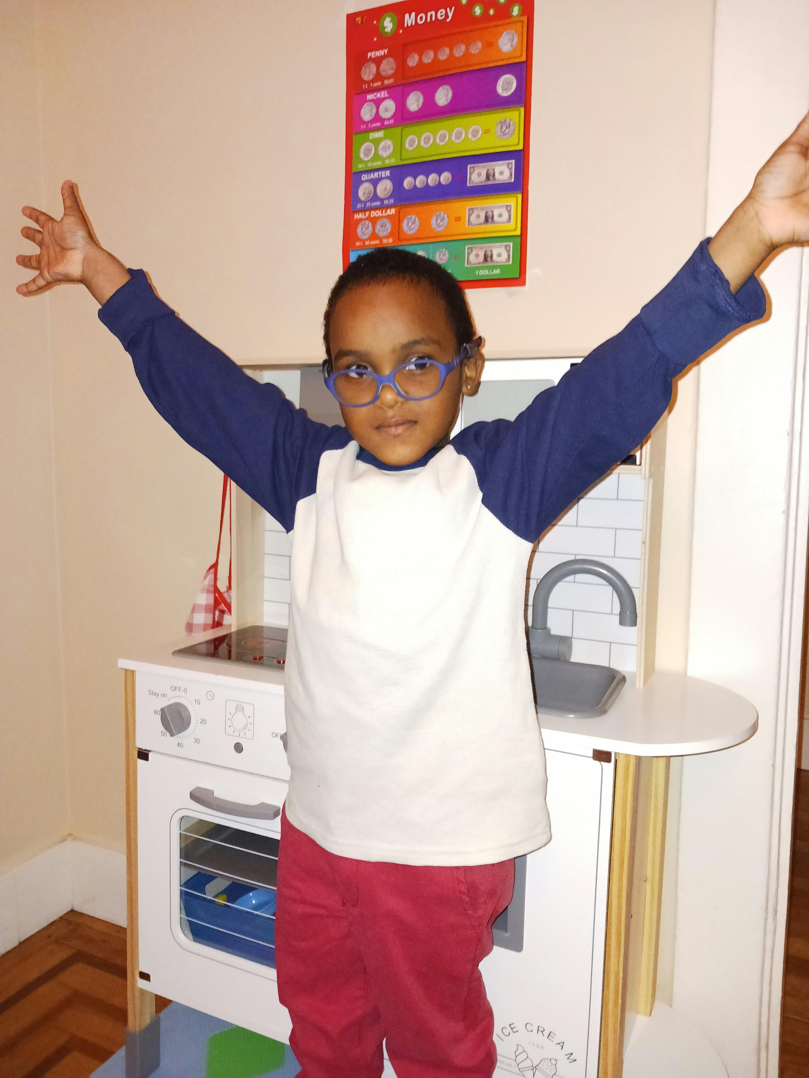 Child with arms raised standing in front of a toy kitchen