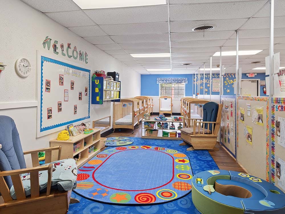 A colorful childcare room with cribs, play mats, and a whiteboard with "WELCOME" above it