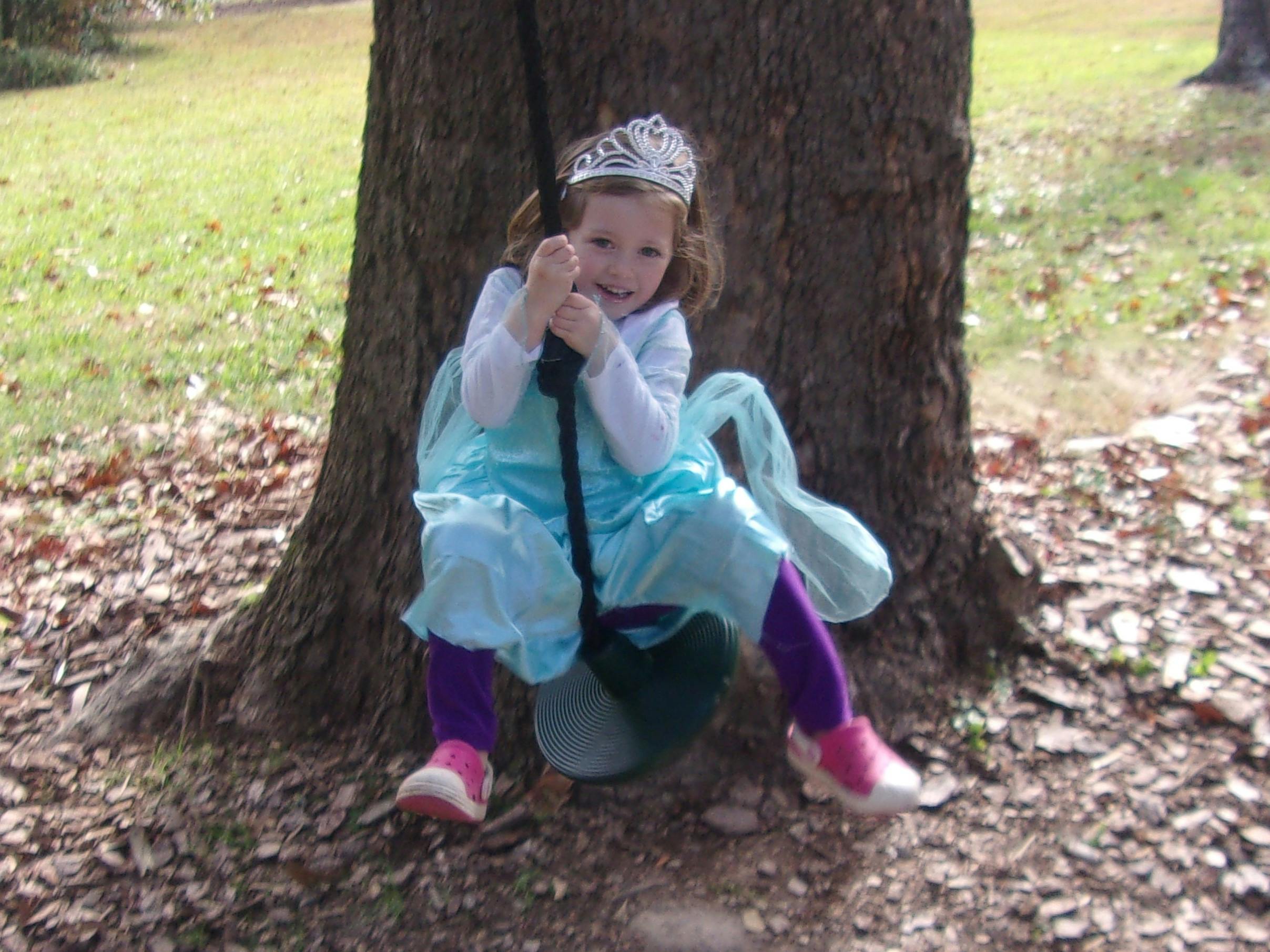 Child in a tiara and costume sitting on a swing attached to a tree