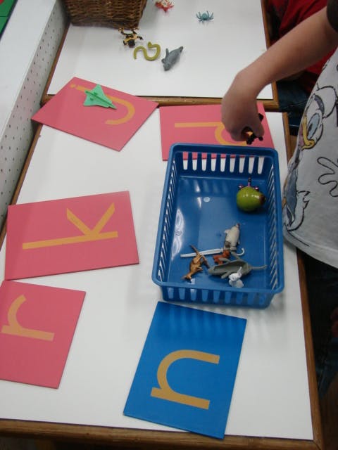 Child's hand reaching for small toys on a table with letter cards