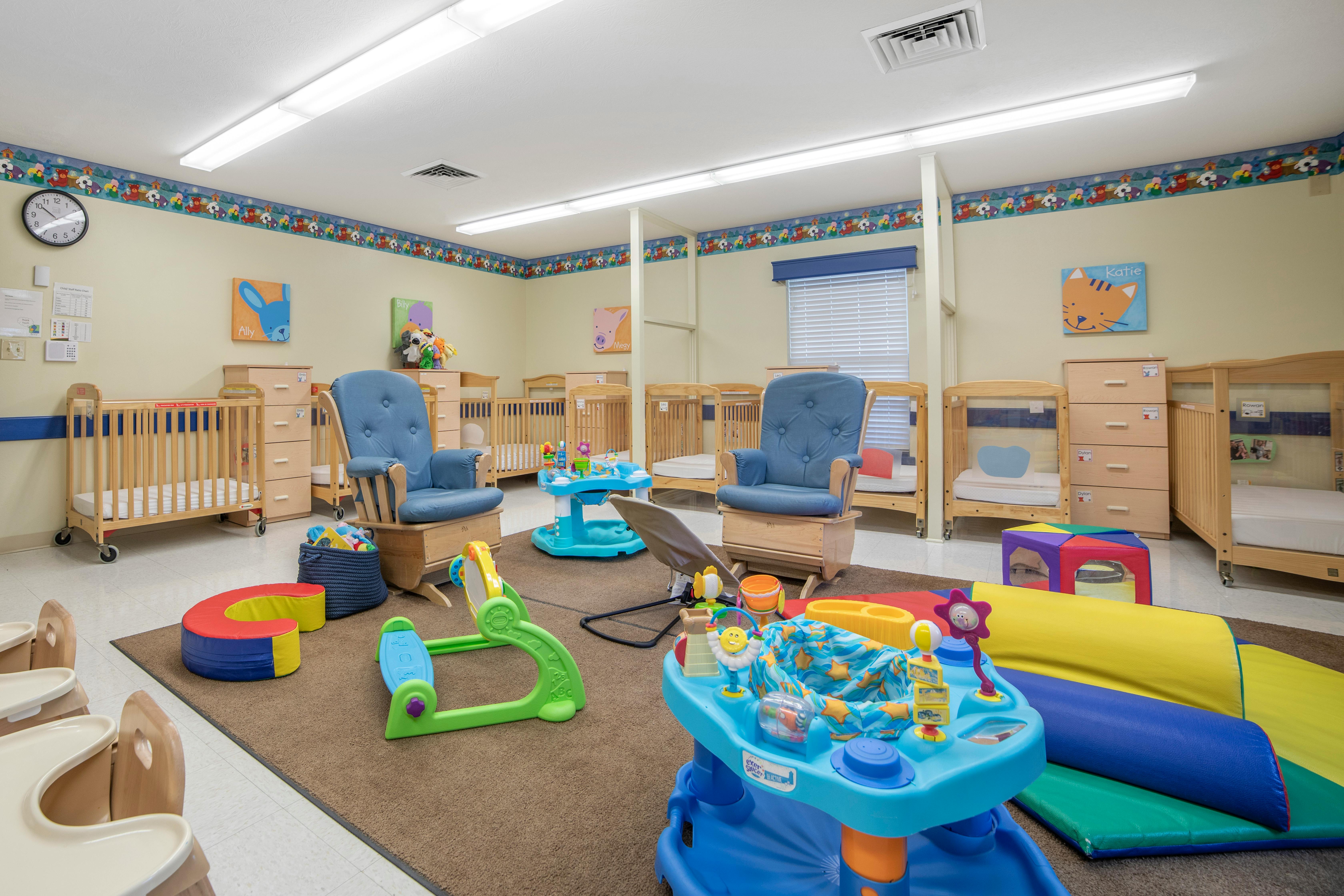 A childcare room with cribs, rocking chairs, and colorful baby toys on a rug