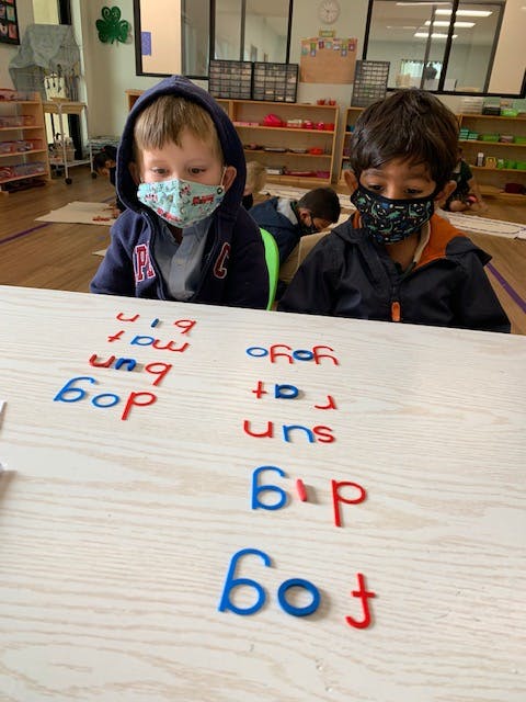 Two children in masks at a table with letter cutouts forming words
