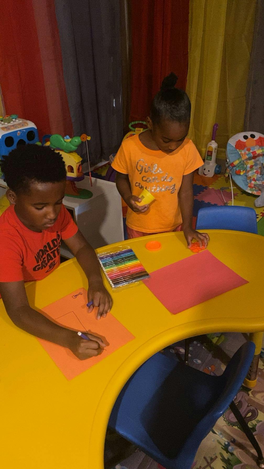 Two children drawing and holding items at a yellow table