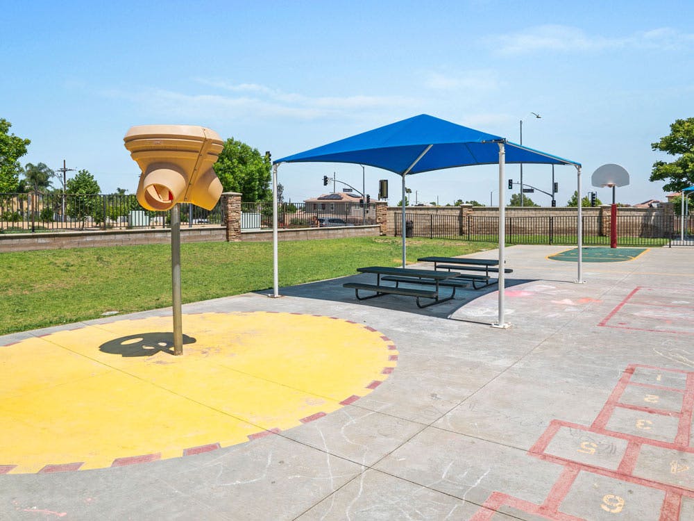 A splash pad with a blue canopy, picnic tables, and a hopscotch court