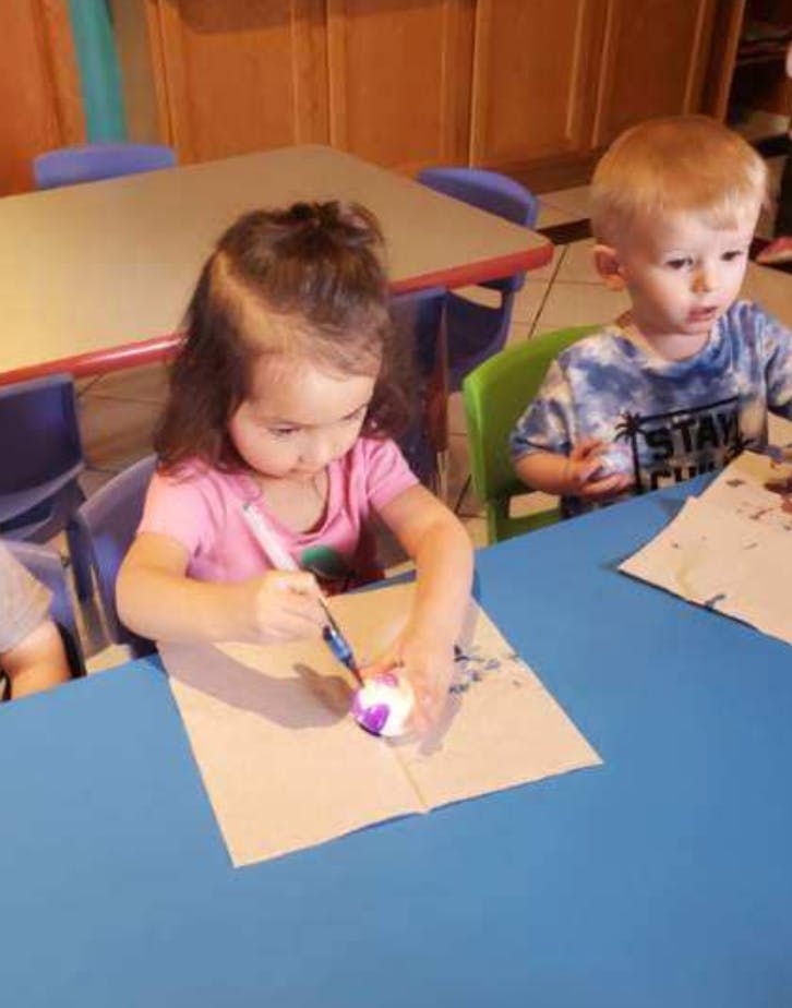 Child painting an object at a table with another child nearby