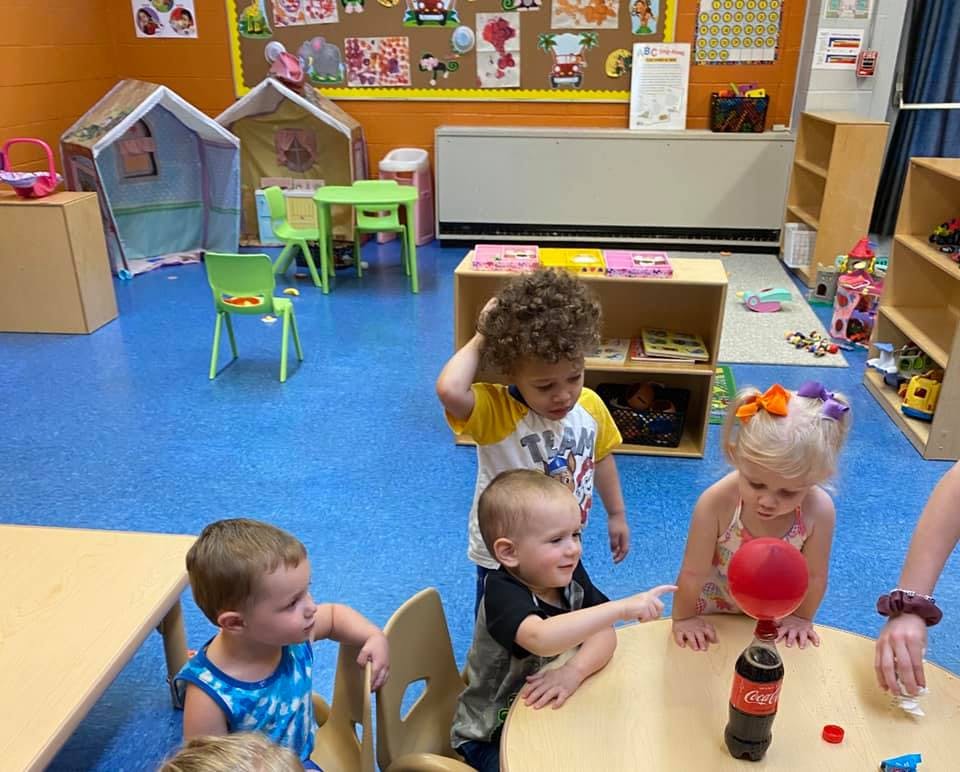 Children playing with a balloon and bottle at a table in a classroom