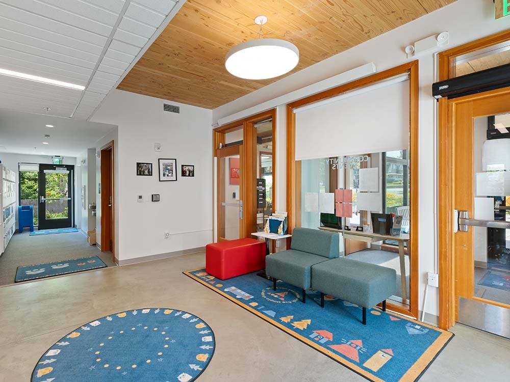 Lobby area with blue rugs, chairs, a red ottoman, and a glass wall to an office