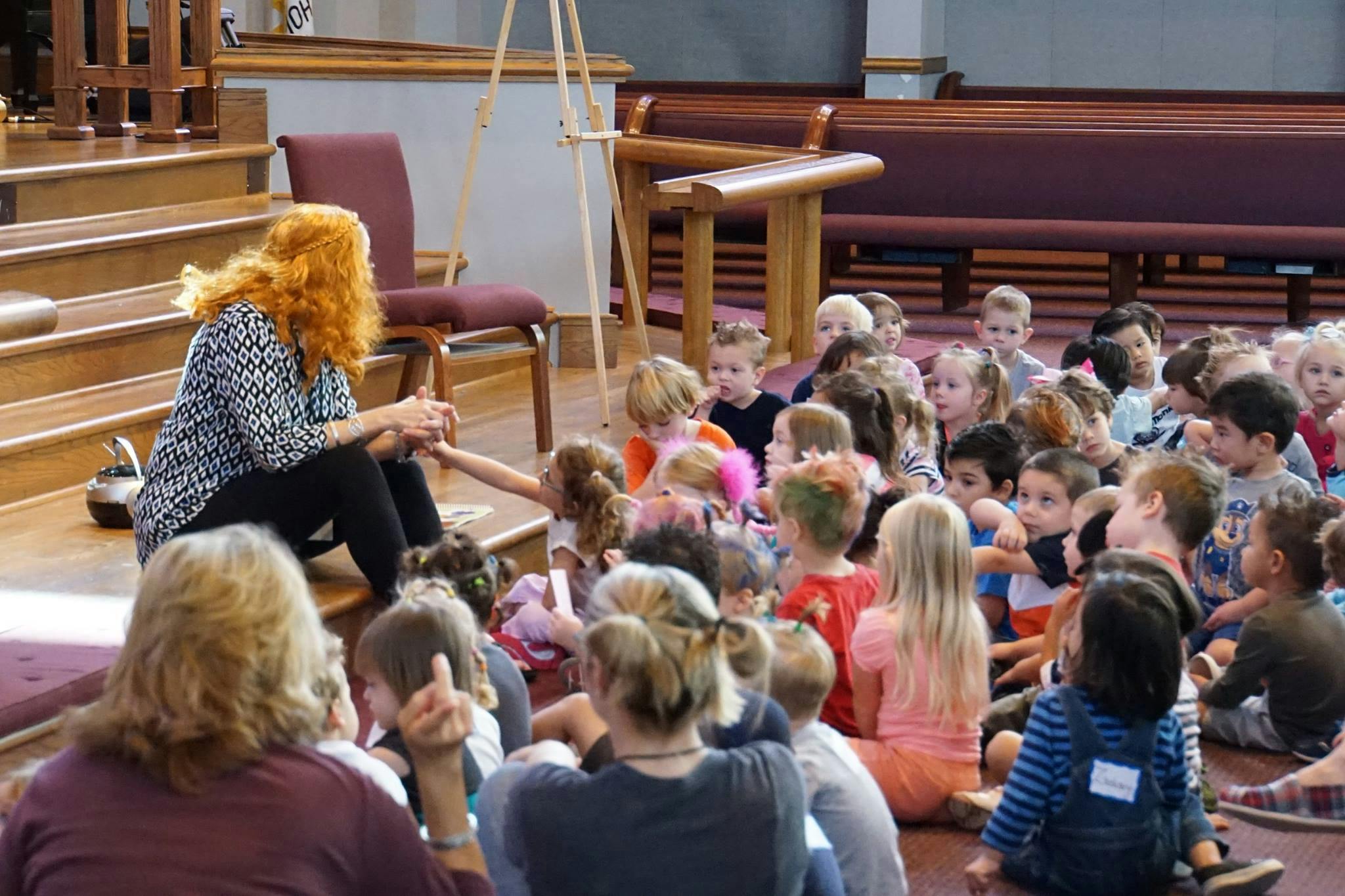 Person with red hair speaking to a group of children sitting on the floor