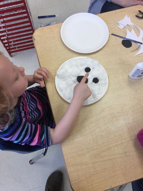 Child pointing at a paper plate craft with cotton and black circles on a table