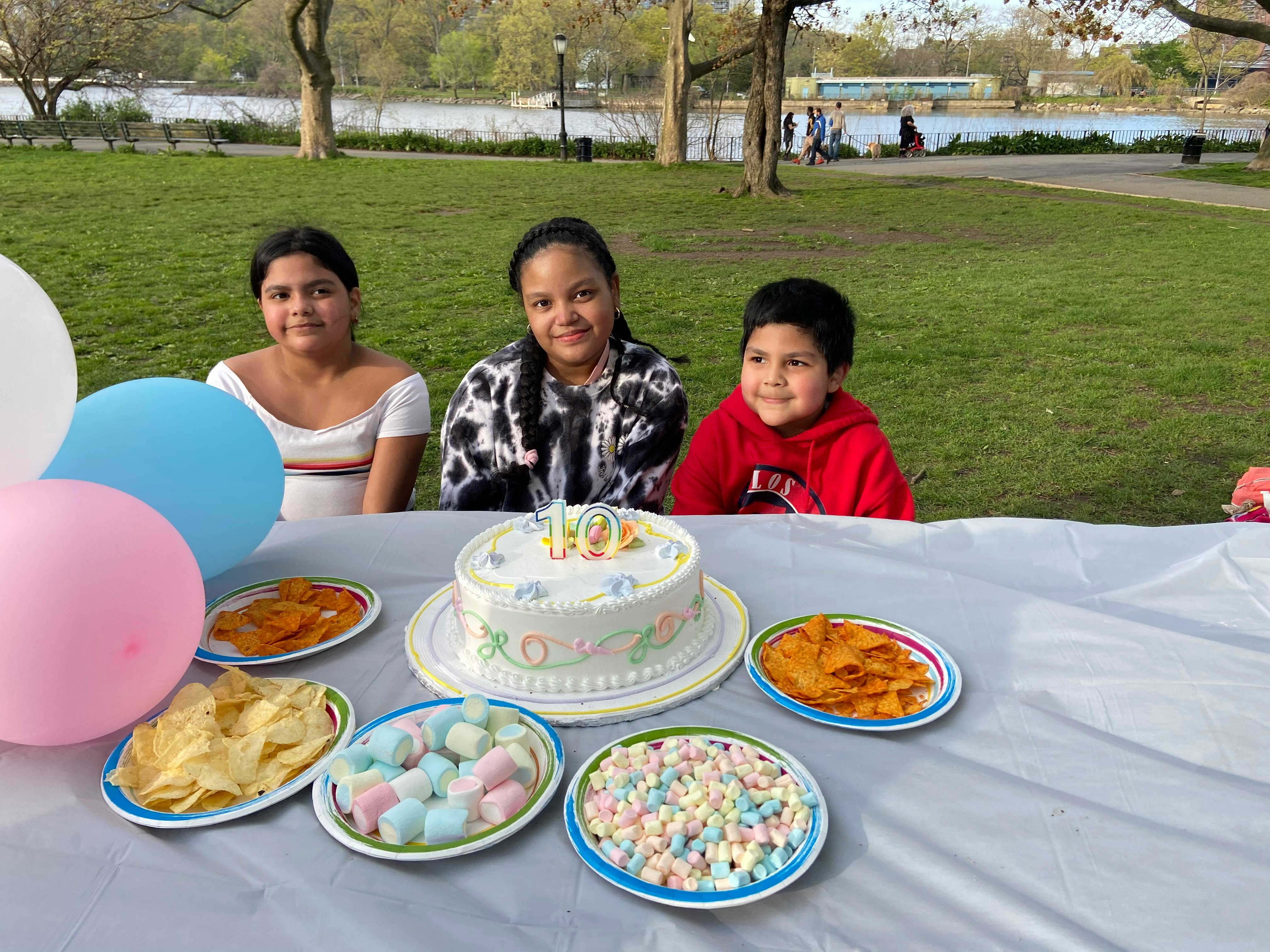 Three people sitting at a table with a birthday cake and snacks outdoors