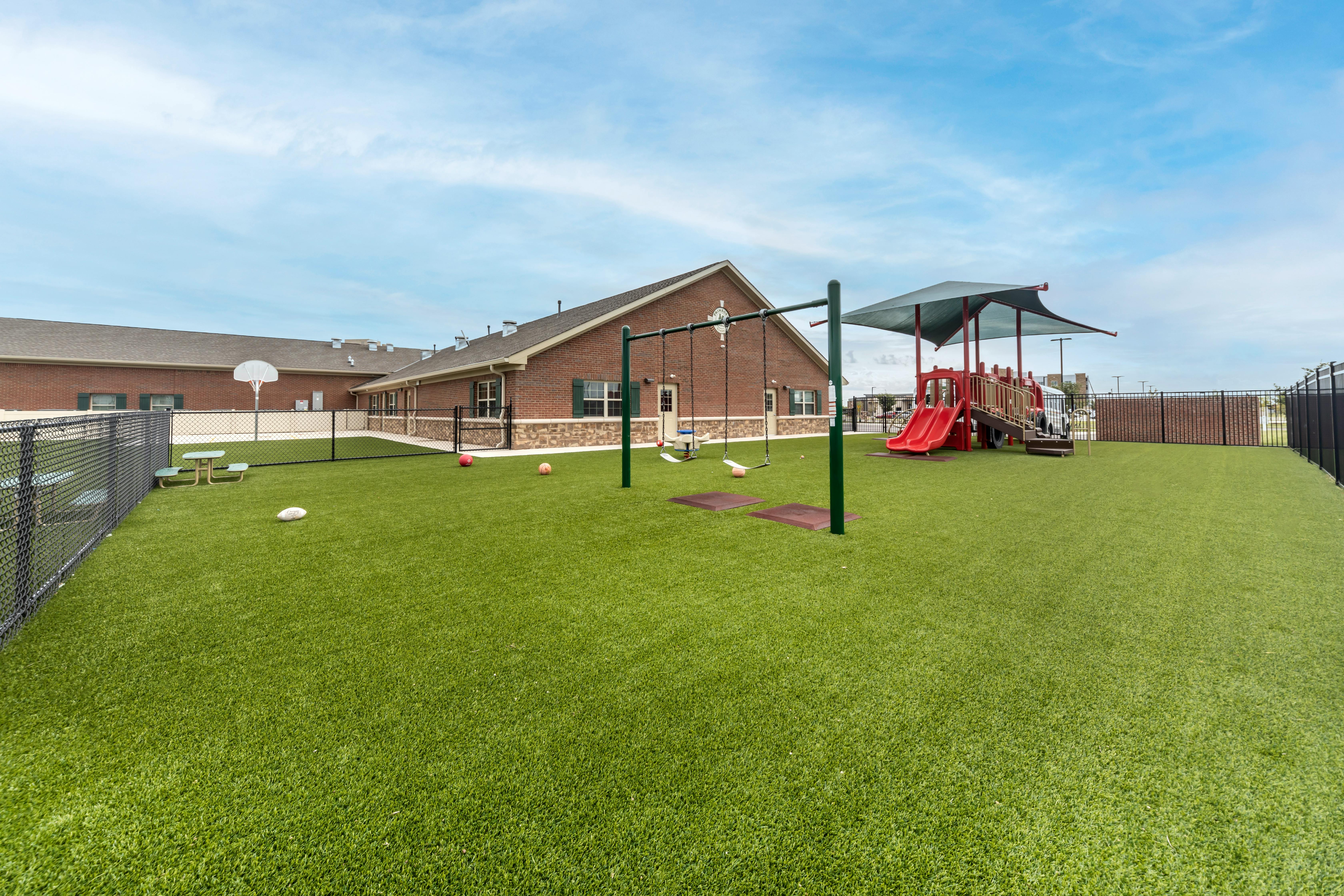 Outdoor playground with green turf, swings, a red slide structure, and a brick building