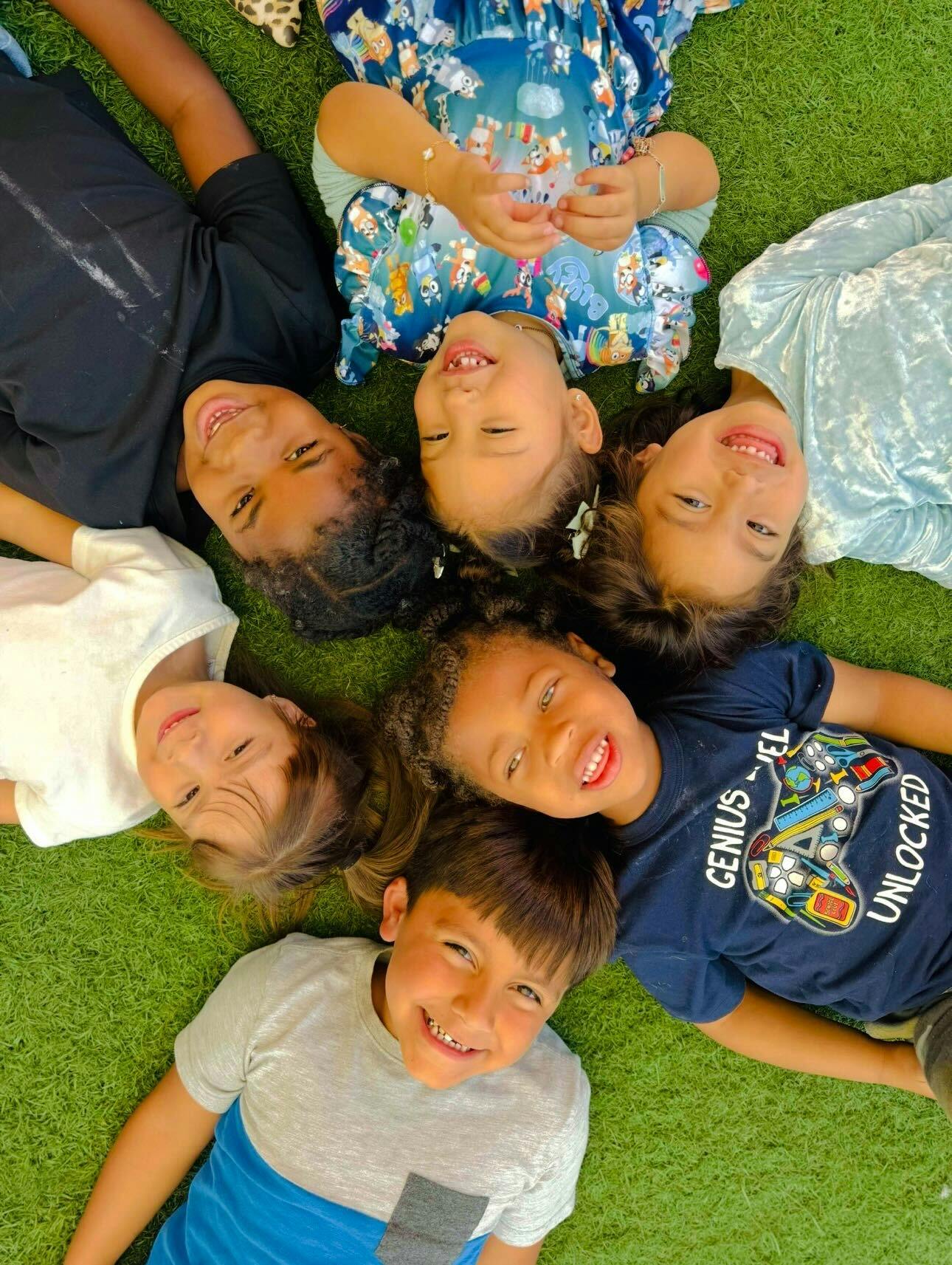 Group of six children lying in a circle on grass, smiling at the camera