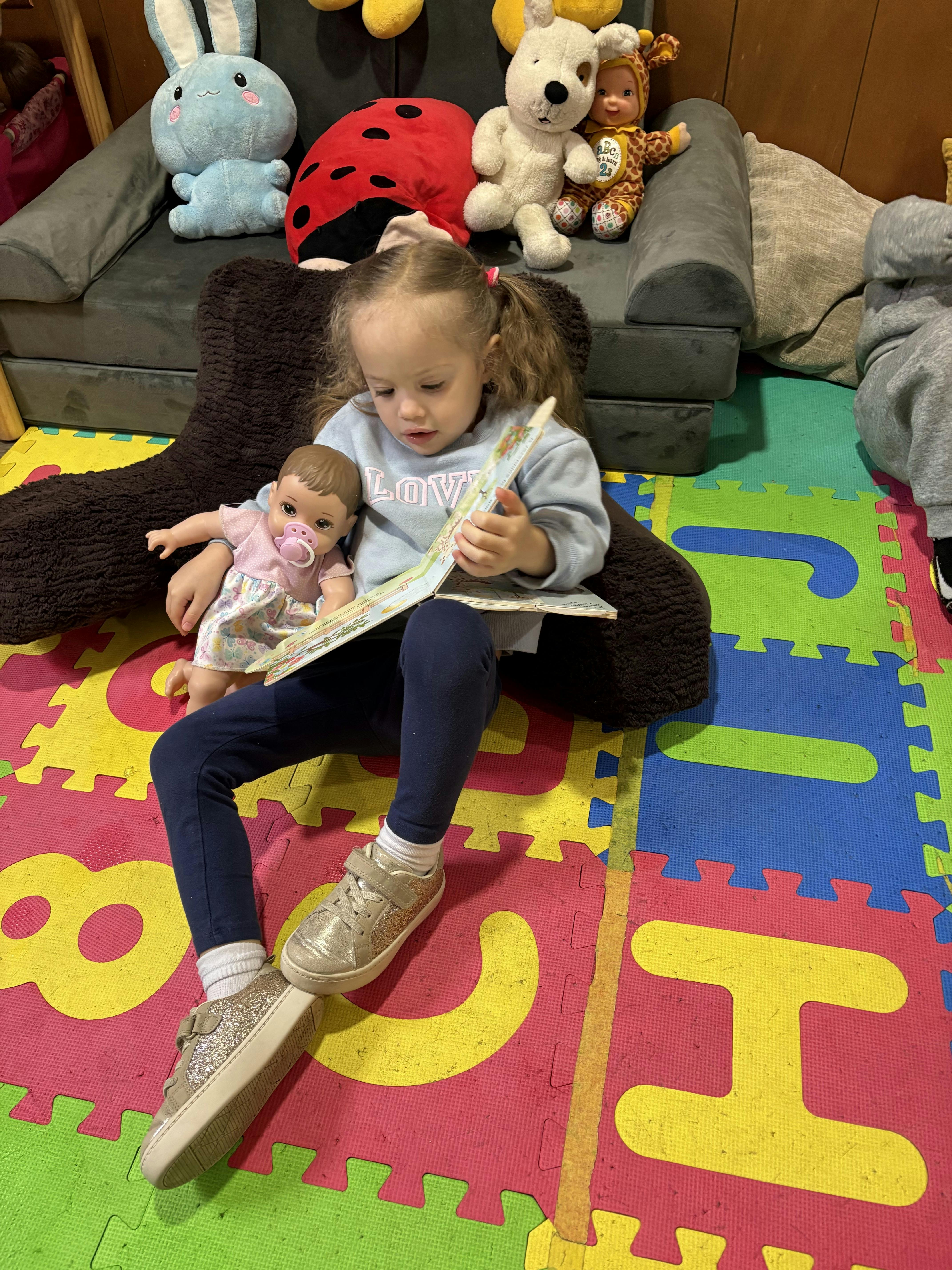 Child sitting on a floor reading a book while holding a doll