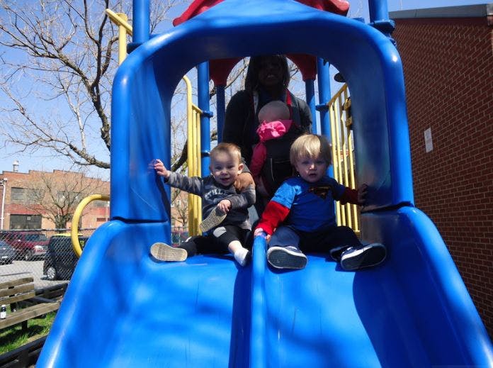 Two children sitting on a blue playground slide with a person holding a baby behind them