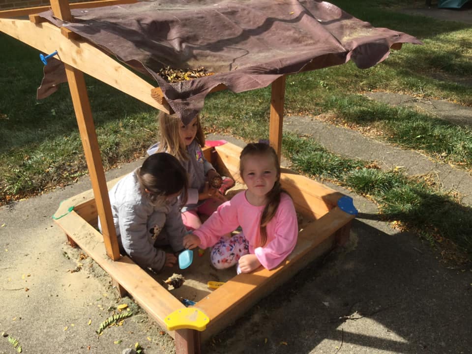 Three children playing in a wooden sandbox with a canopy