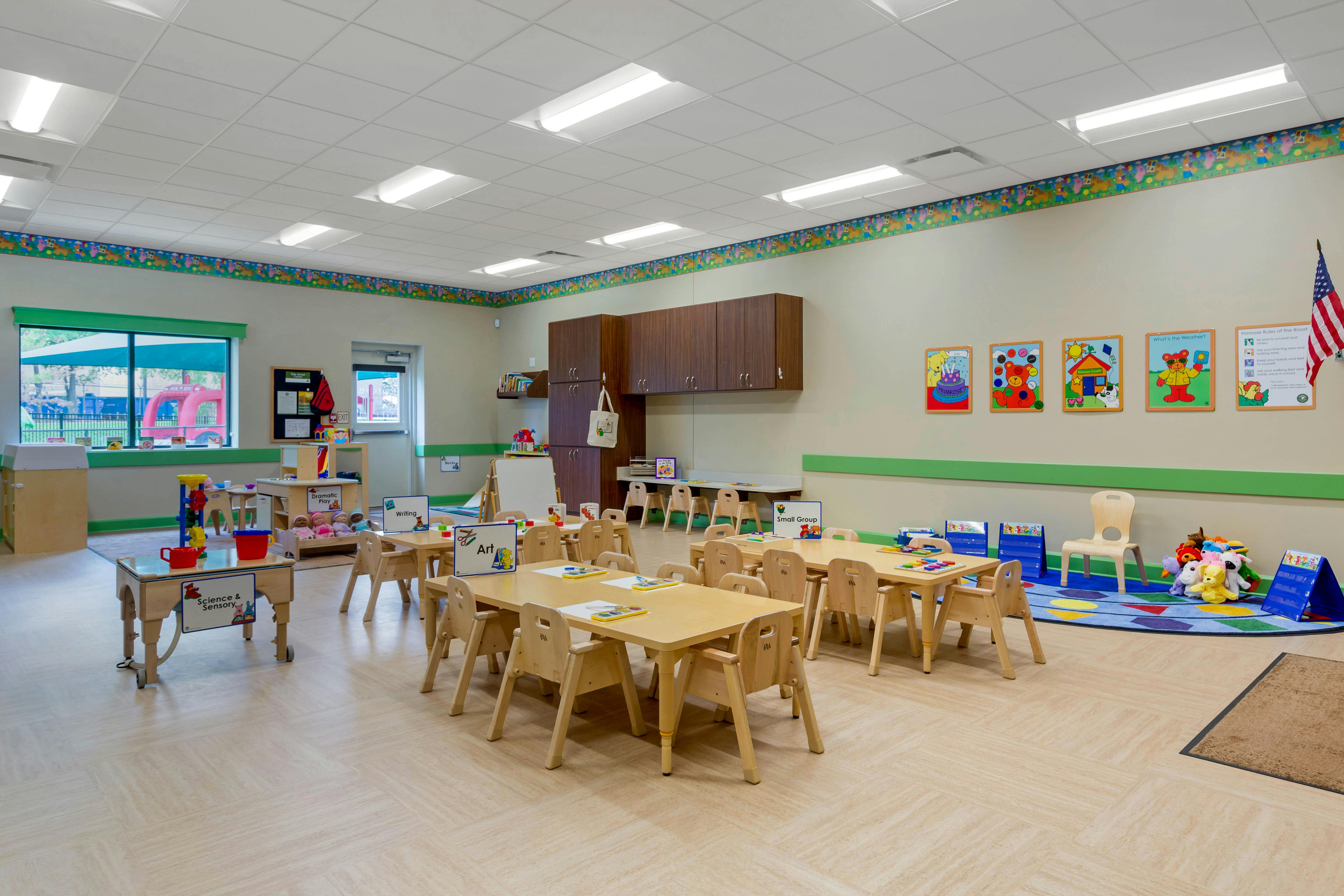 Classroom with child-sized tables and chairs, learning stations, and a rug with toys