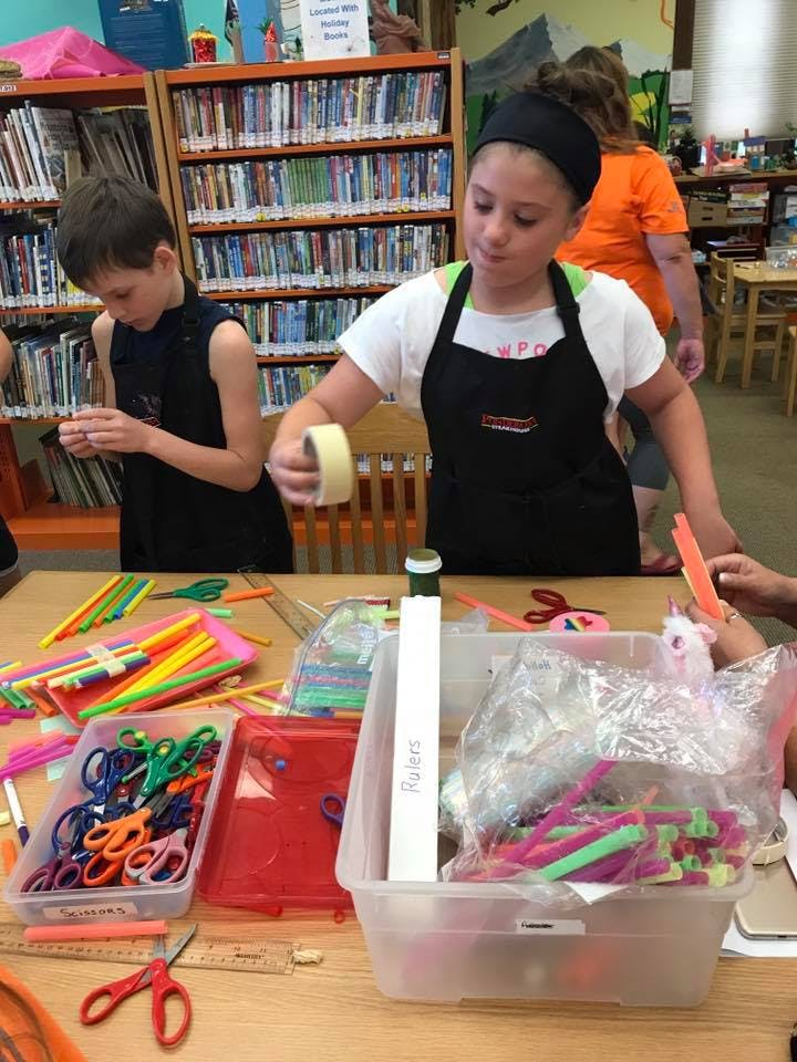Child in apron holding tape, another child working on a craft project