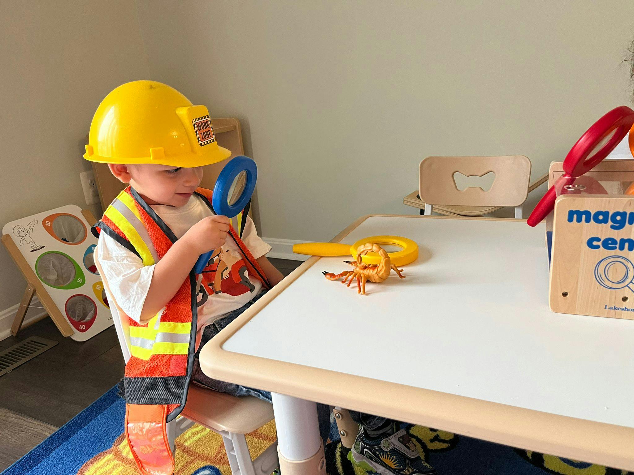 Child wearing a construction hat and safety vest looking at a toy scorpion with a magnifying glass