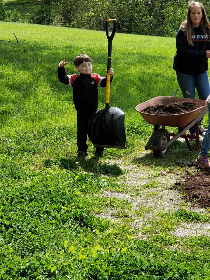 Child holding a large shovel next to a wheelbarrow of dirt