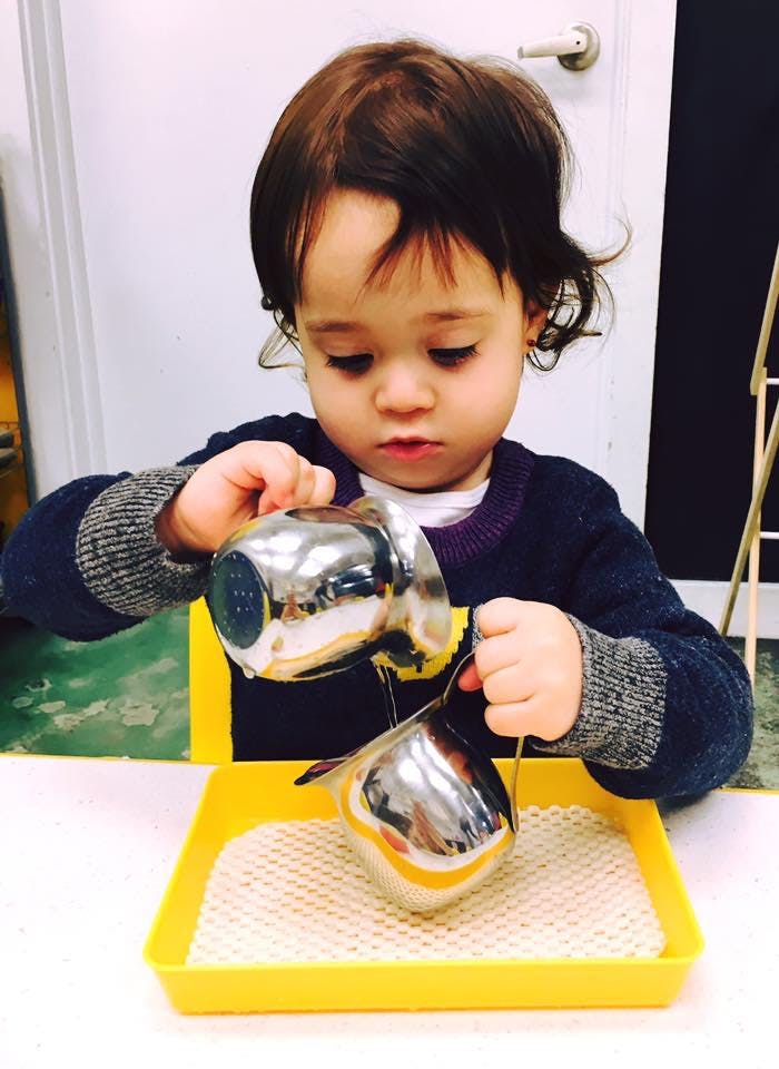 Child pouring from one metal pitcher into another over a yellow tray