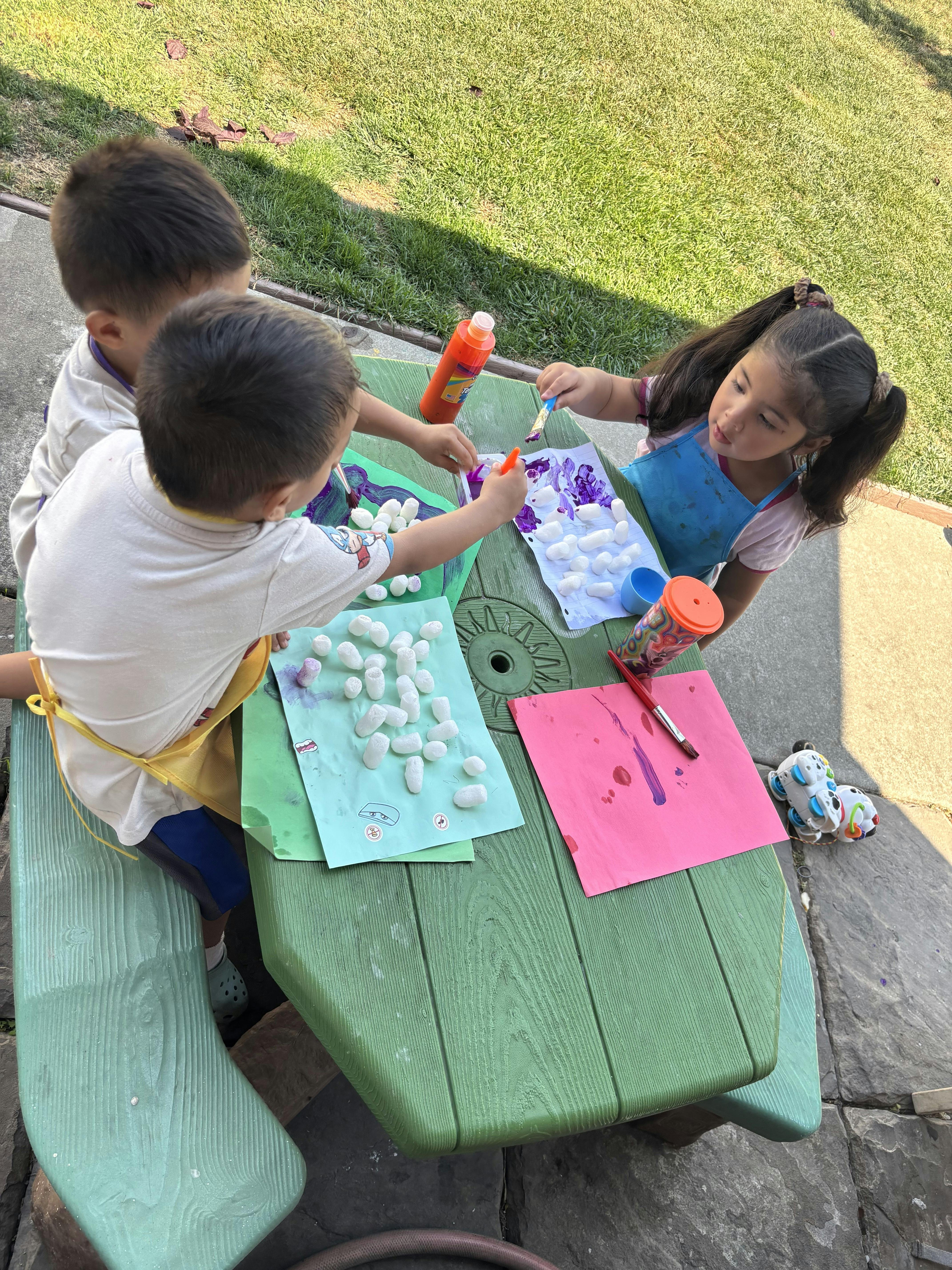 Three children painting small white objects at a green picnic table outdoors
